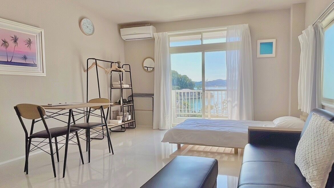 A modern hotel room with a bed, a sofa, a desk with chairs, a balcony with a view of the sea, and a painting on the wall.