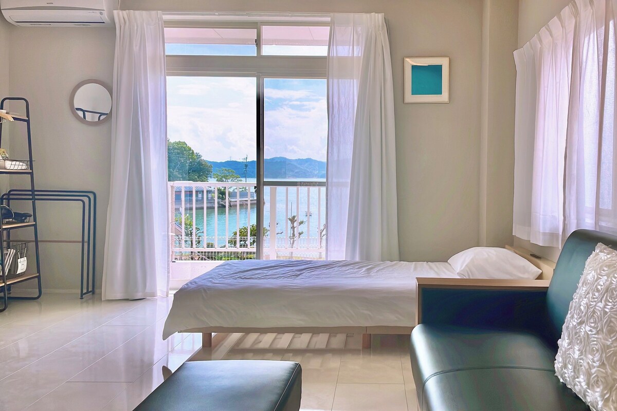 A room with a bed, a sofa, a small table, a shelf with items, and a view of a body of water and mountains.