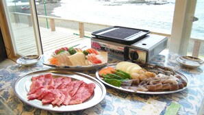 Mesa com diversos alimentos e uma churrasqueira, vista de um quarto com janela para a praia.