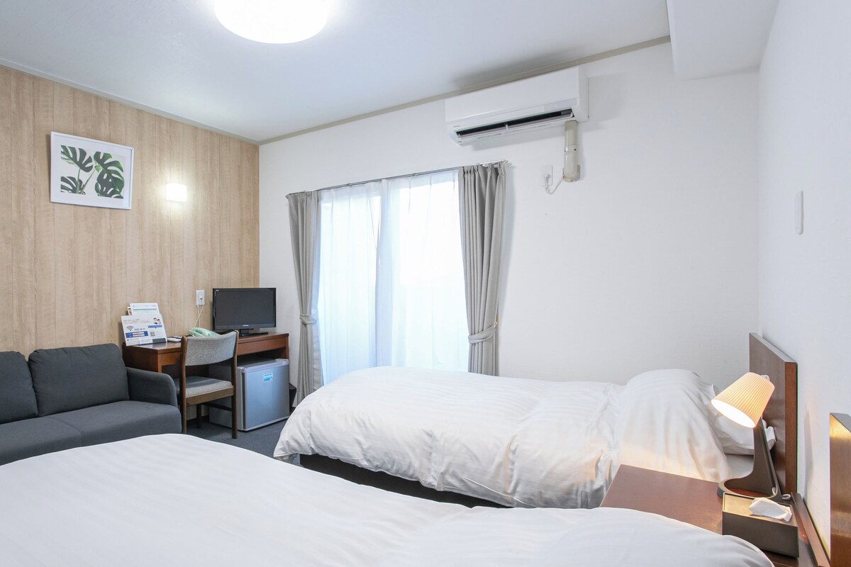 Habitación de hotel con dos camas, un escritorio con computadora, un sofá y una ventana con cortinas.