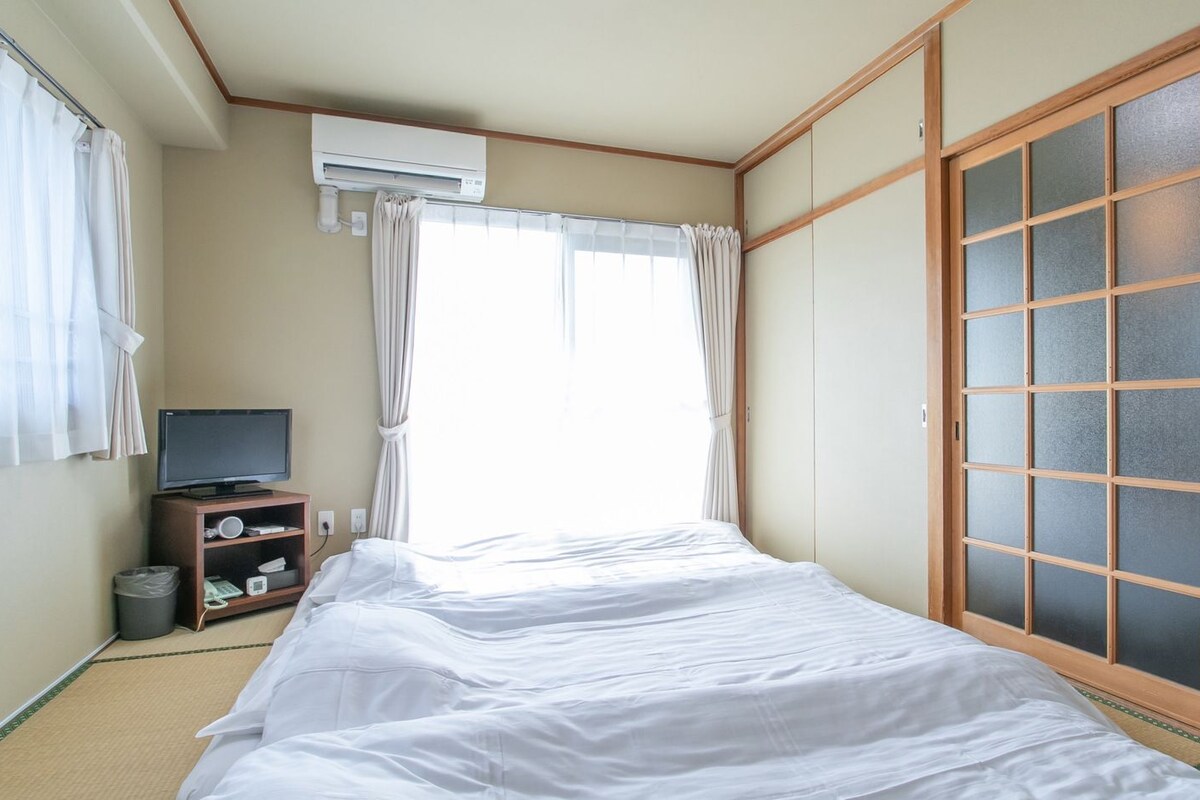 Una habitación con una cama, un televisor sobre un mueble, una ventana con cortinas y una puerta corredera.