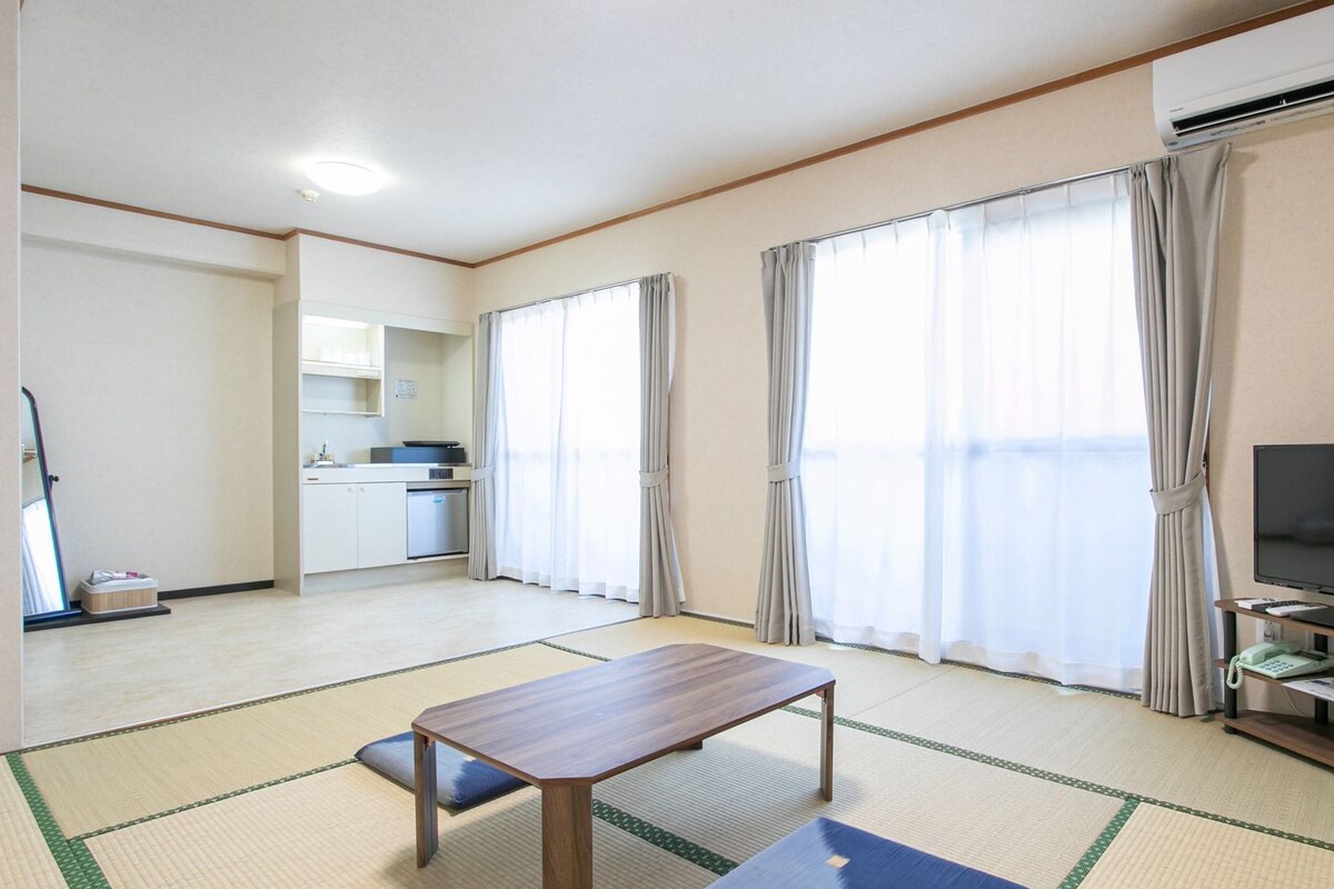 Una habitación con suelo de tatami, un televisor sobre un mueble bajo, una mesita y una zona de cocina al fondo.
