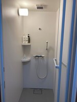 Un espacio de ducha pequeño y cerrado, con ducha de mano, luz empotrada en la pared y estante con artículos de aseo.