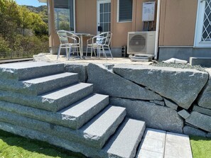 Escalones de piedra exteriores que llevan a un edificio con puerta de cristal, una mesa con sillas y un equipo de aire acondicionado.