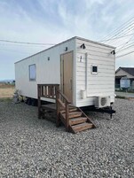 Una vivienda móvil de una sola planta, pequeña, con una escalera de madera que lleva a una puerta, un ventanal y un equipo de aire acondicionado.