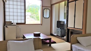 Una habitación japonesa tradicional con suelo de tatami, una mesa baja, un sofá, un televisor y una puerta corredera que da a un balcón.
