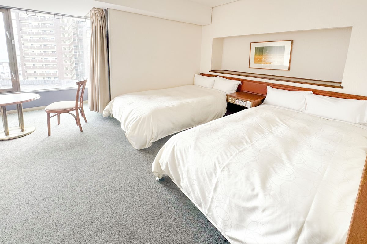 Ein Hotelzimmer mit zwei Einzelbetten, einem Holznachttisch, einem kleinen Tisch mit Stuhl und einem großen Fenster mit Vorhängen.