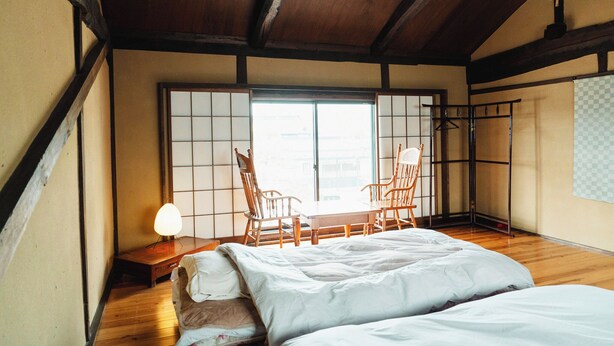 A traditional Japanese room with tatami flooring, a low wooden bed, a table with chairs, and a sliding door. - Idemaru Guest House (Ueda)