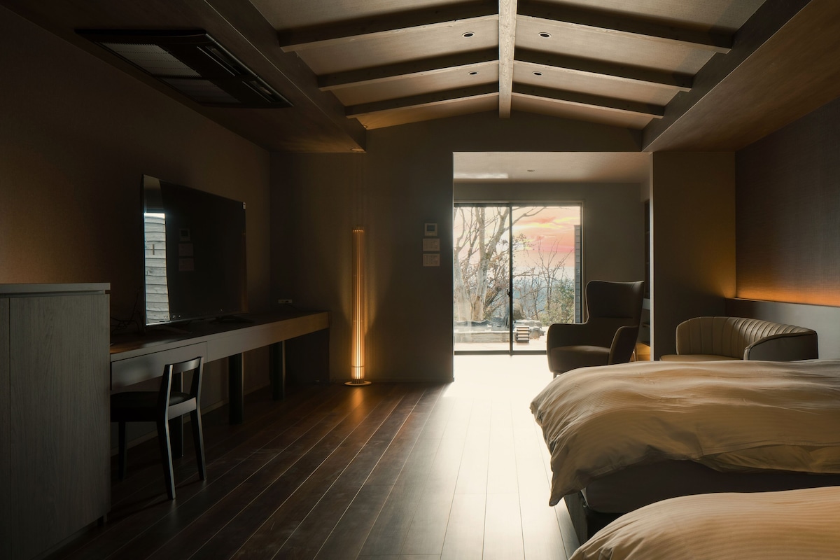 Un dormitorio moderno con una cama grande, un escritorio y una silla. Se ve una vista de árboles a través de un ventanal.