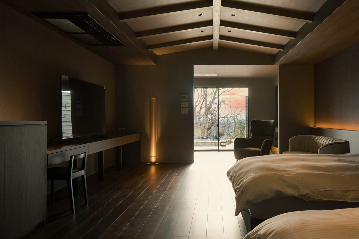 Een moderne slaapkamer met een groot bed, een bureau en een stoel. Er is uitzicht op bomen door een glazen deur.