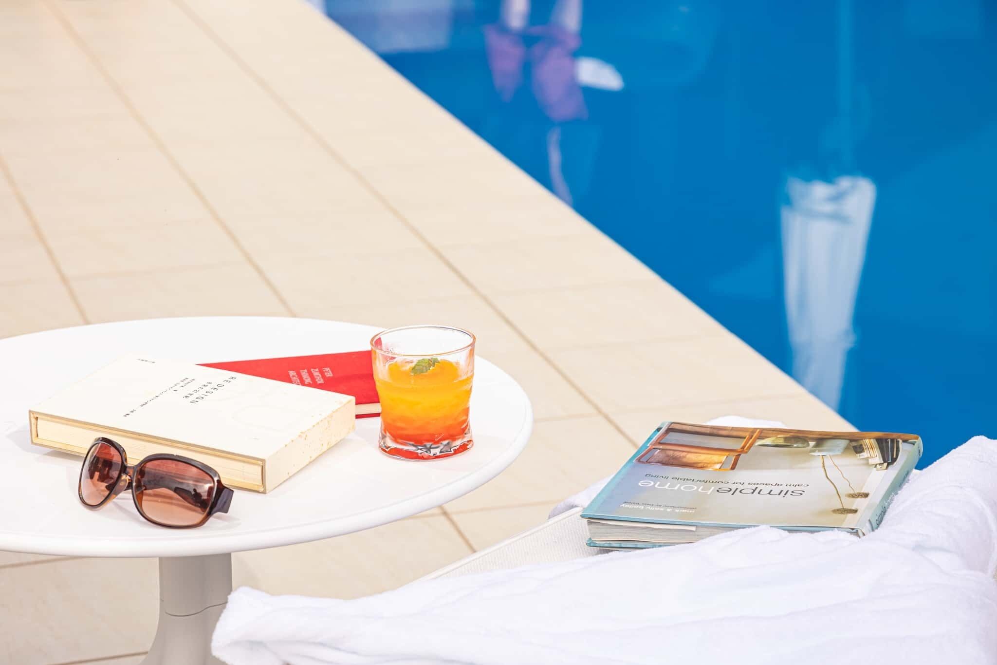 A poolside table with a book, a pair of sunglasses, a glass of drink, and a magazine.