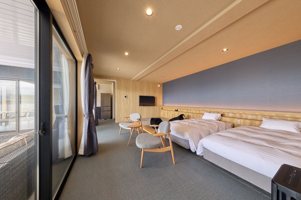 A hotel room with two beds, a desk, a chair, a TV, and a large window.