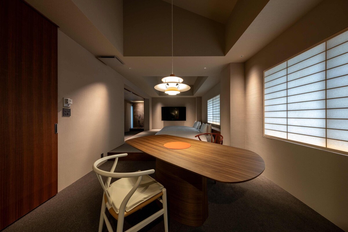 Quarto de hotel moderno com uma escrivaninha de madeira curva, uma cama de solteiro, uma cadeira e uma janela grande com persianas.