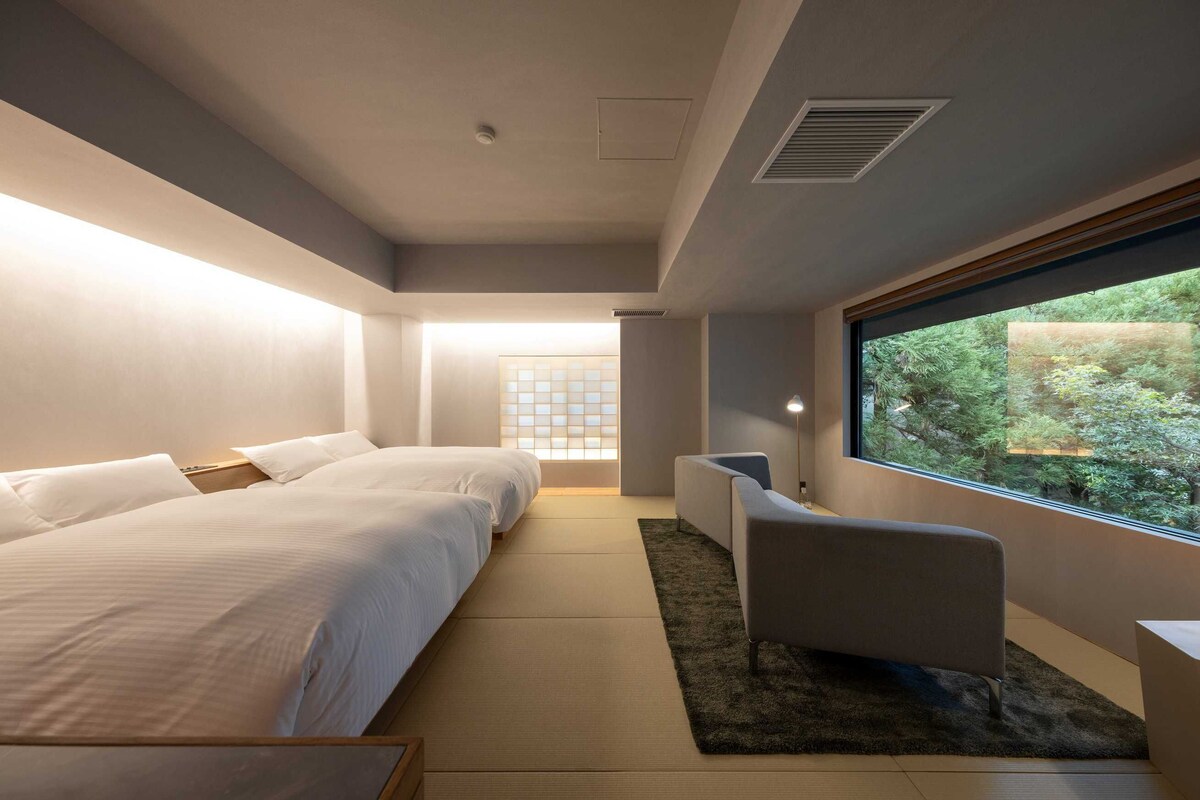 Quarto de hotel moderno com duas camas, área de estar e uma grande janela com vista para o verde.