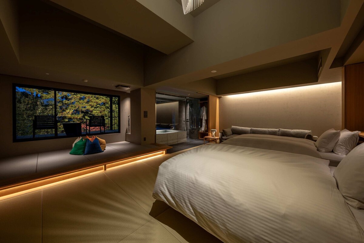Quarto moderno com uma cama grande, área de estar com almofadas, vista para um jardim através de uma janela ampla e uma banheira integrada.
