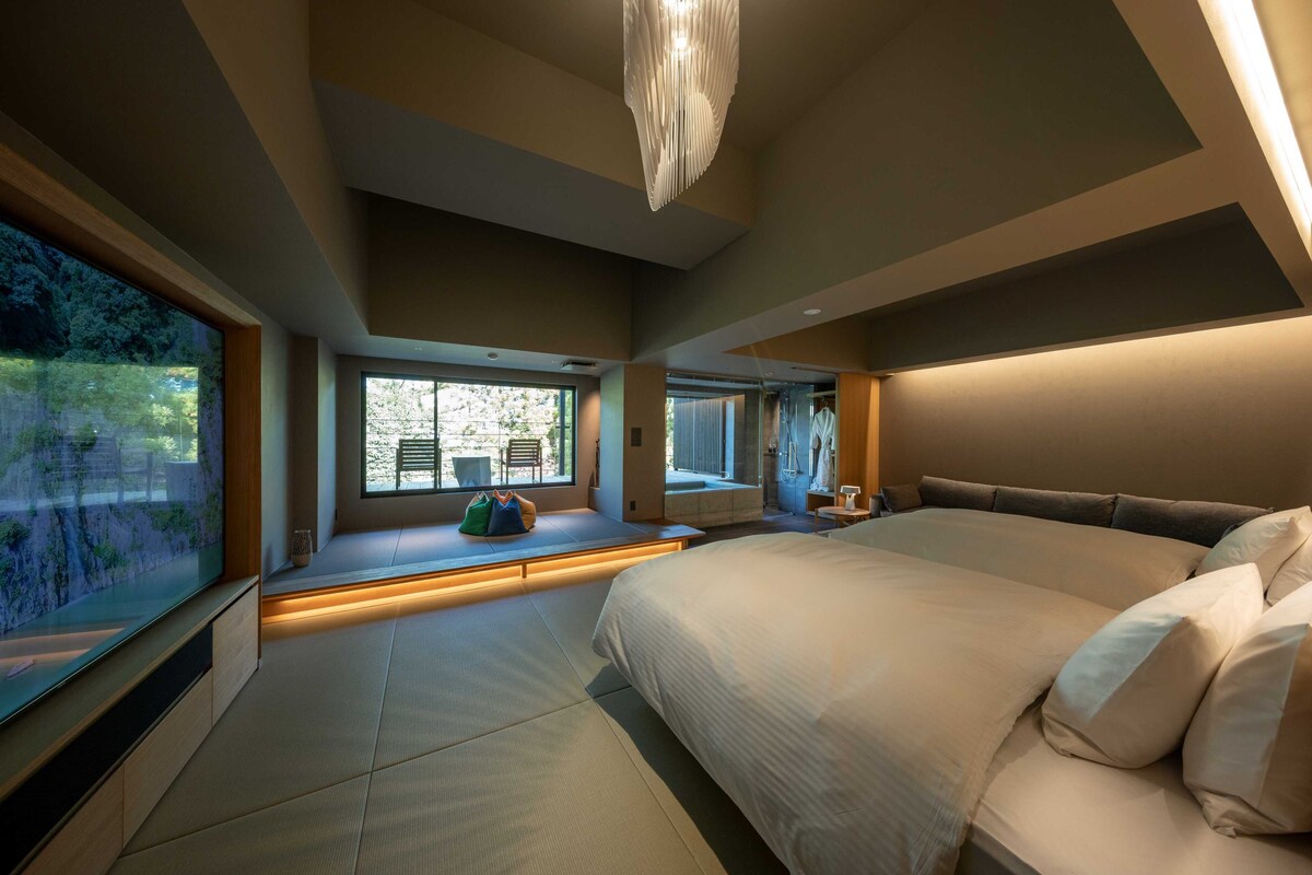 Quarto moderno com cama grande, área de estar integrada e amplas janelas.