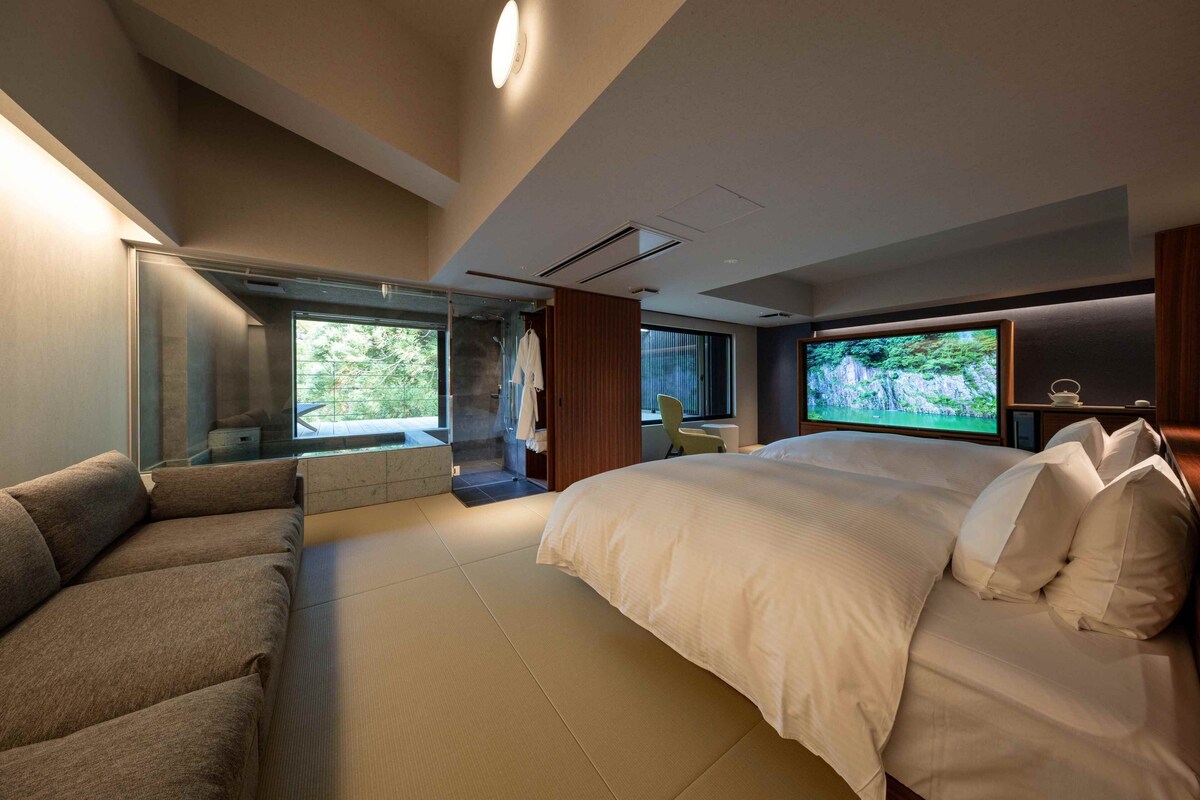 Quarto de hotel moderno com uma cama grande, um sofá, uma televisão e vista para o verde lá fora.