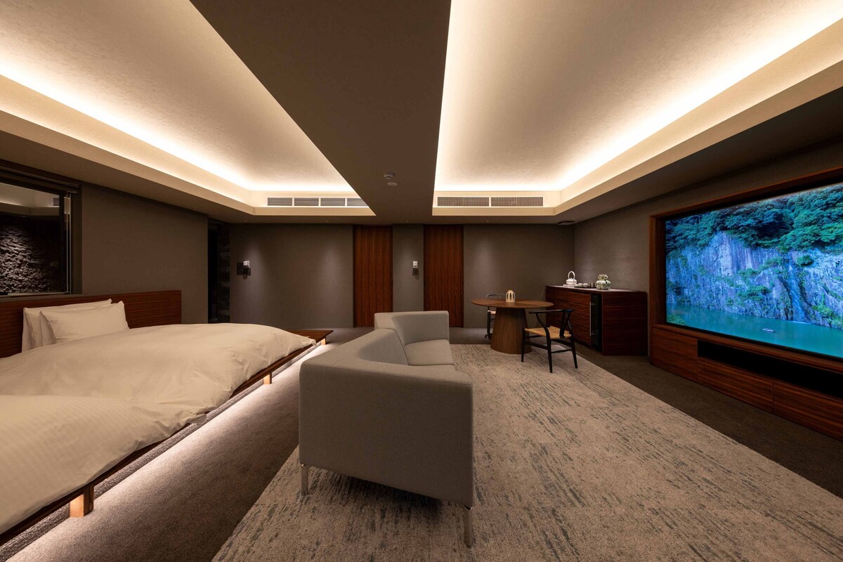 Quarto de hotel com uma cama grande, um sofá, uma televisão e uma escrivaninha com uma cadeira.