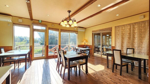 Un comedor con piso de madera, mesas de vidrio y sillas, ventanas amplias con cortinas y un candelabro.
