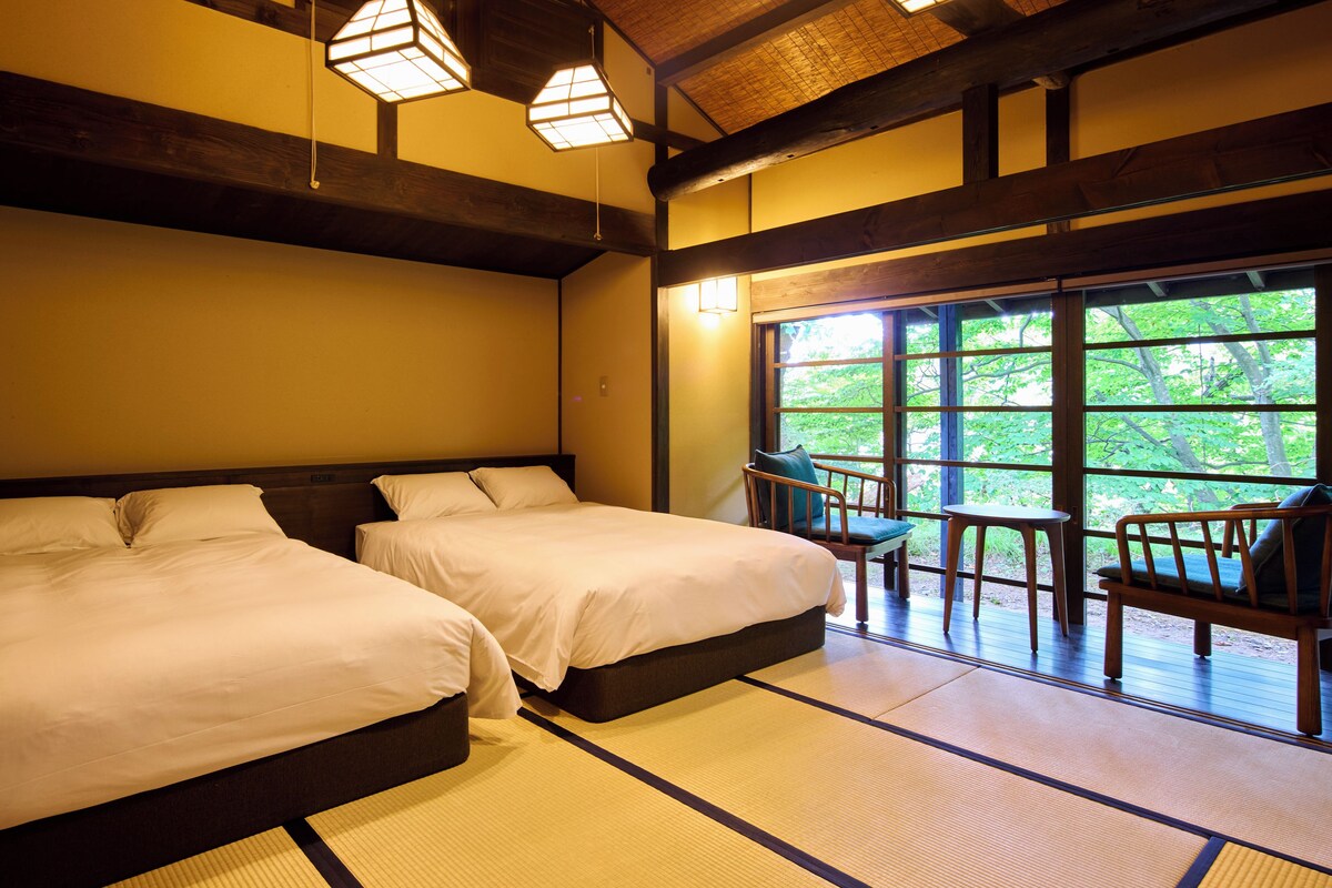 Een traditionele Japanse kamer met tatami vloeren, twee bedden, een houten plafond en uitzicht op groen door grote ramen.