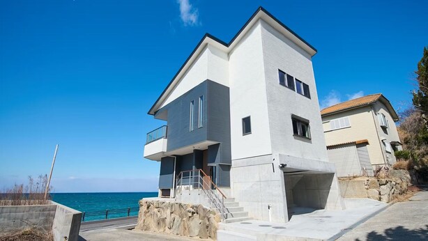 Ein modernes, zweistöckiges Haus mit Balkon, einer Steinsichtmauer und Meerblick.