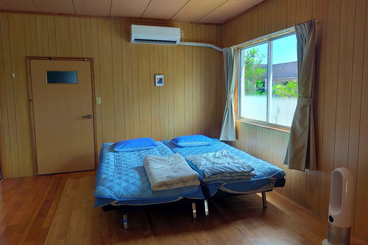 A room with wooden panel walls, a bed with blue bedding, a window with curtains, and an air conditioning unit.