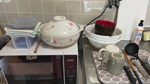 A kitchen with a microwave, stacked towels, a floral pot, a red bowl, a mug, and utensils on a tray.