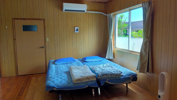 A room with wooden panel walls, a bed with blue bedding, a window with curtains, and an air conditioning unit.