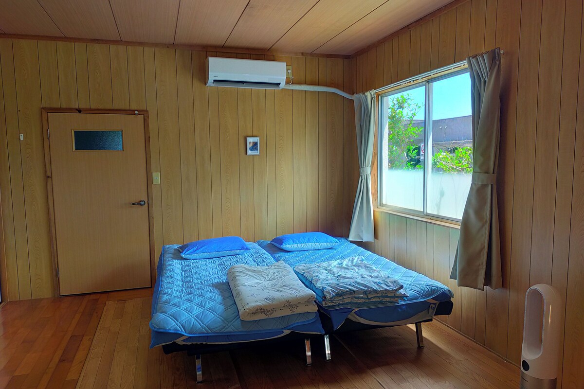 Una habitación con paredes de madera, una cama con ropa de cama azul, una ventana con cortinas y un equipo de aire acondicionado.