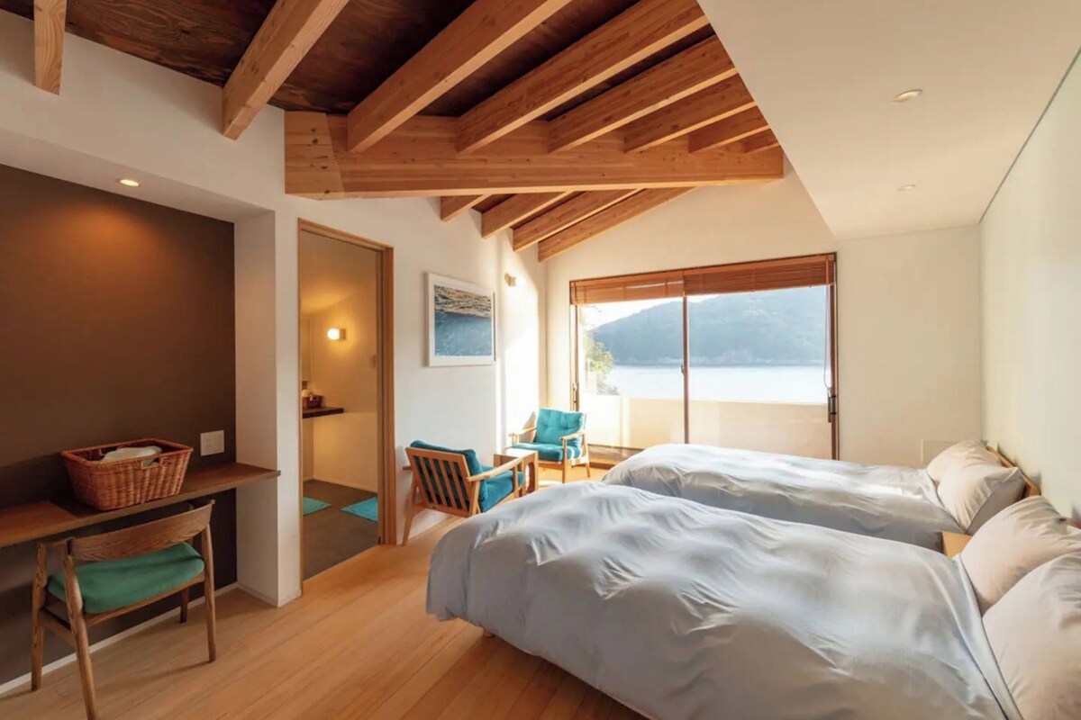 A bedroom with two beds, a wooden ceiling, a window with a view, and a small desk with a chair.