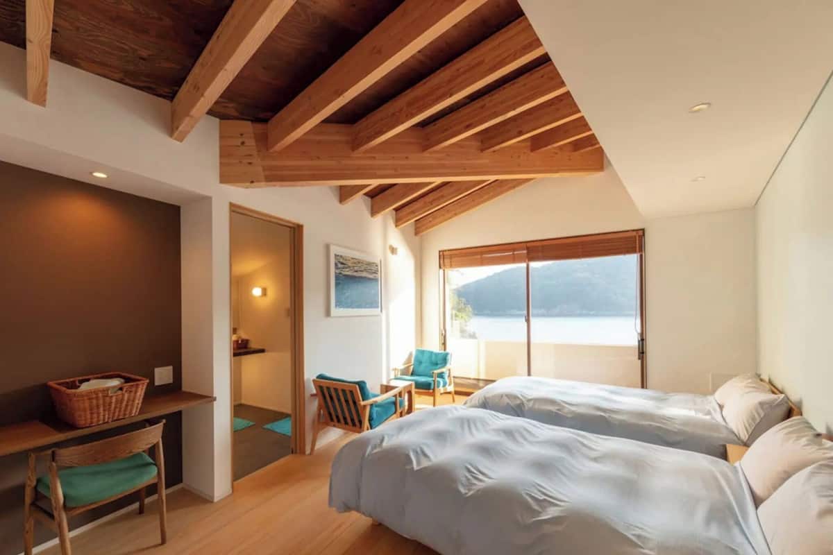 A bedroom with two beds, a wooden ceiling, a window with a view, and a small desk with a chair.