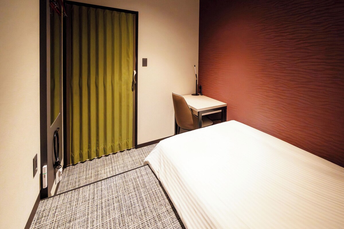 Una camera d'albergo con un letto, una scrivania, una sedia e una tenda verde.