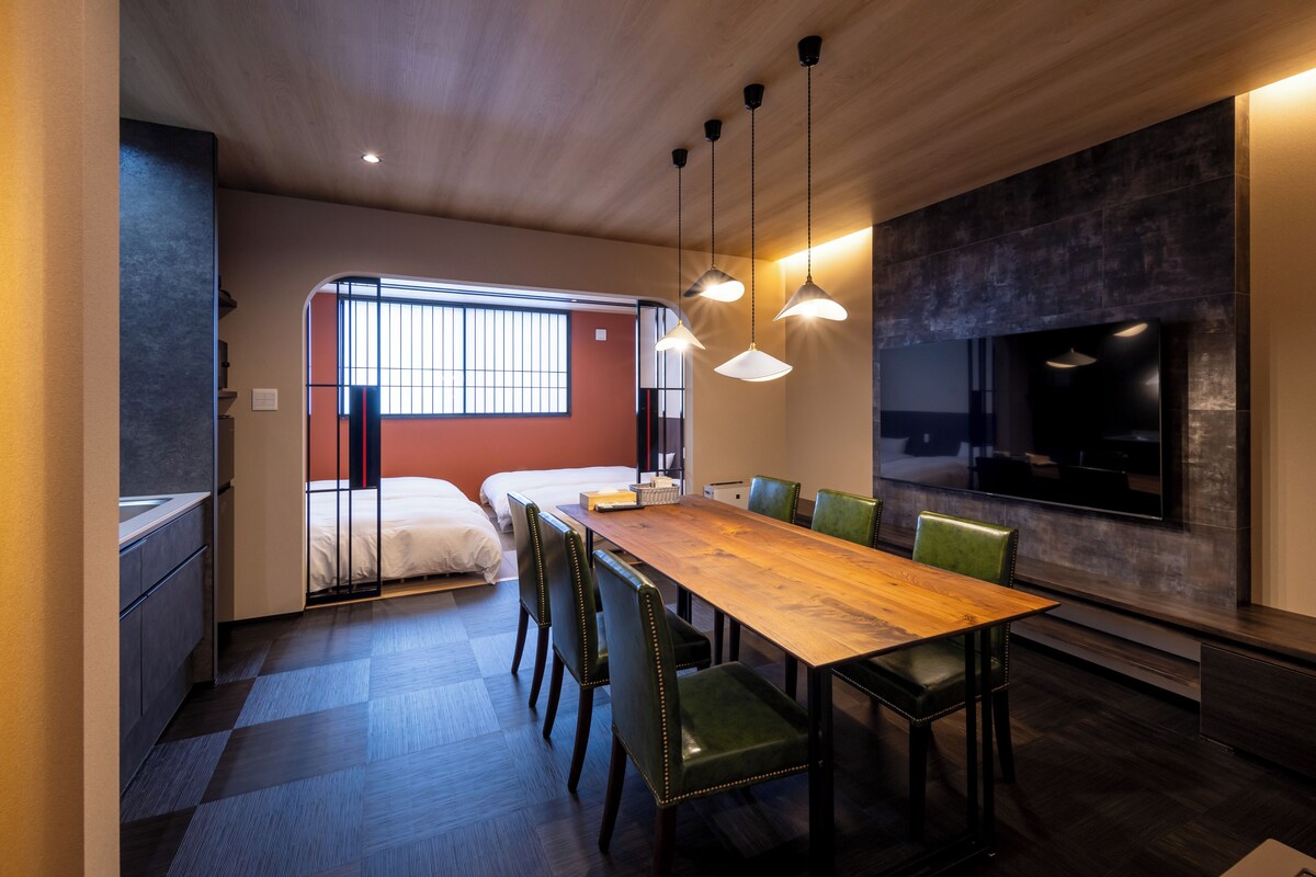 Een moderne hotelkamer met een houten eettafel, groene beklede stoelen, een grote televisie aan de muur en een bed met een rode accentmuur.