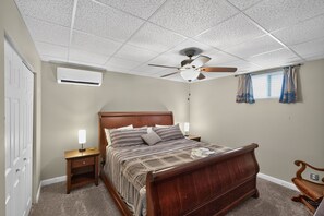 Ein Schlafzimmer mit einem hölzernen Bett, einem Deckenventilator, einer Klimaanlage, einem Nachttisch mit Lampe und einem Fenster mit Vorhängen.
