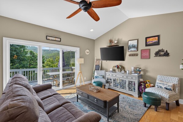 A spacious living room with a large sofa, a coffee table, and a television. There is a balcony with a view of trees and a mountain. - Blue Ridge Retreat | Hot Tub, Game Room & Views (Fairview)