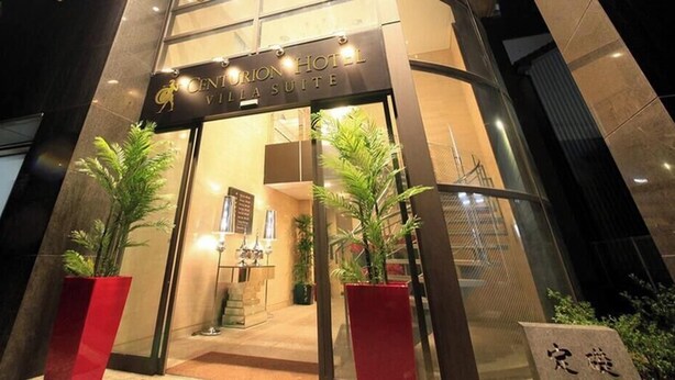 The entrance to Centurion Hotel with a glass facade, potted plants, and a visible staircase leading to the interior.