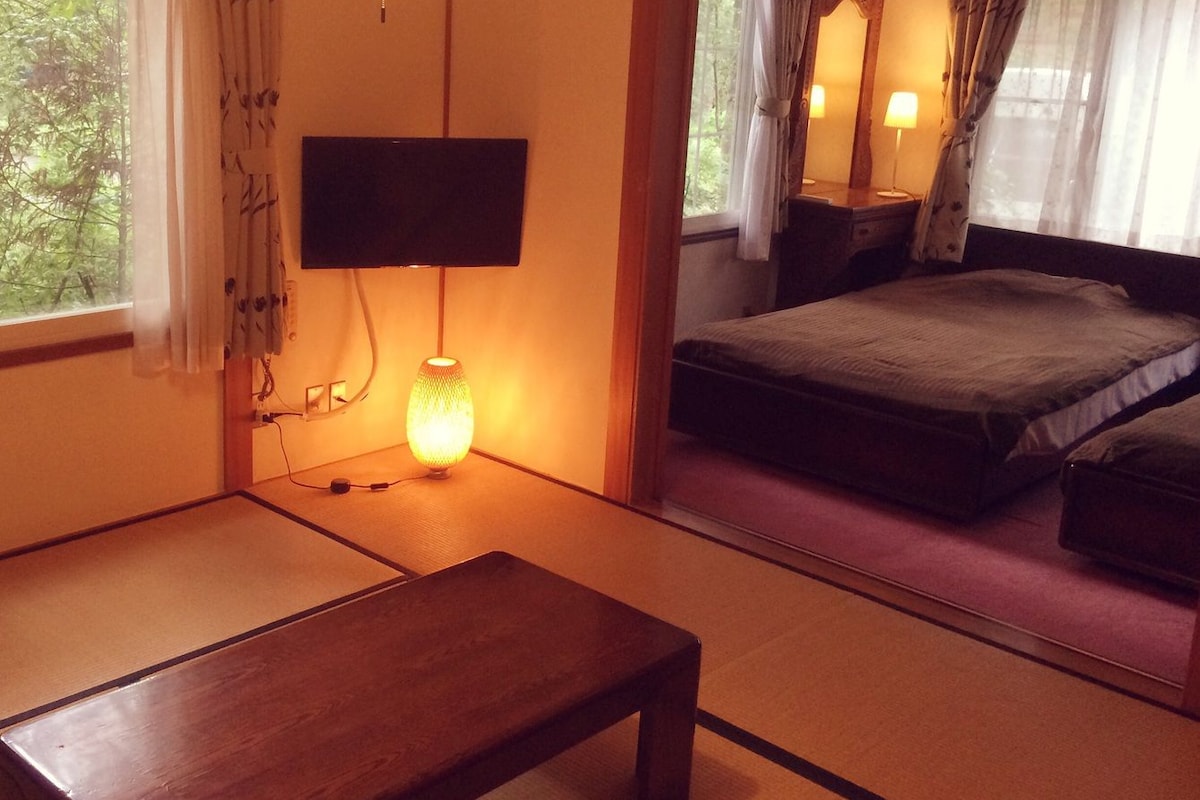 A traditional Japanese-style room with tatami flooring, a low table, a bed with a dark bedspread, a wall-mounted TV, and a bedside lamp.