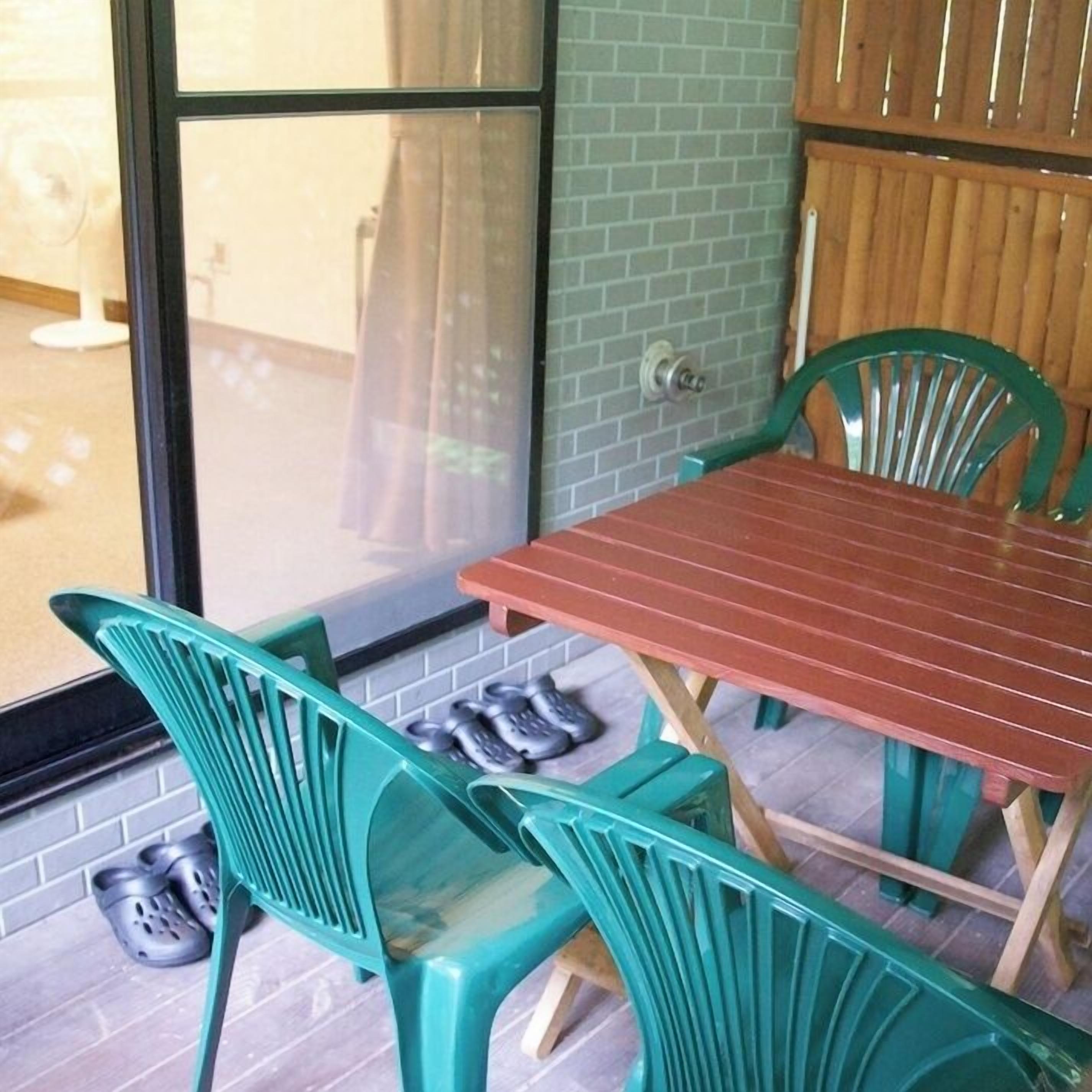 Quarto com mesa de madeira e cadeiras de plástico verde, janela com vista e parede de tijolos.