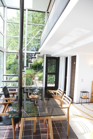 A modern dining area with a glass table and wooden chairs, large windows, and a staircase leading to an outdoor area. - Izu Cliff House (Minamiizu)