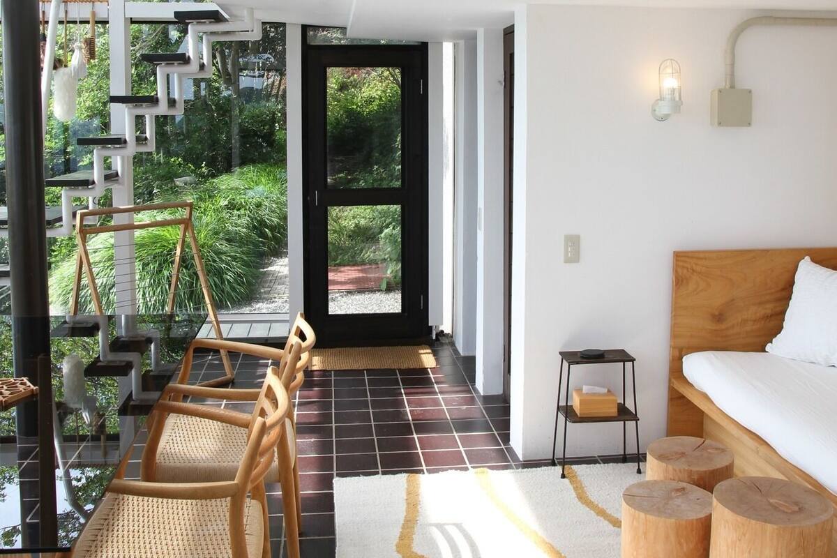 Una habitación de hotel moderna con una cama grande, un cabecero de madera, una mesita de noche pequeña y una silla. Hay una mesa de vidrio con sillas fuera de la habitación y una escalera que lleva a un balcón.