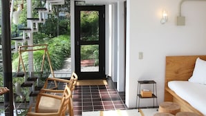Una habitación de hotel moderna con una cama grande, un cabecero de madera, una mesita de noche pequeña y una silla. Hay una mesa de vidrio con sillas fuera de la habitación y una escalera que lleva a un balcón.