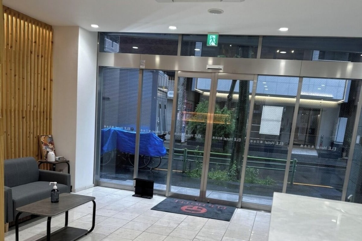 Zona de recepción de hotel con diseño moderno, que incluye un mostrador de recepción, un área de estar con sofá y mesa, y amplias puertas de vidrio que dan al exterior.