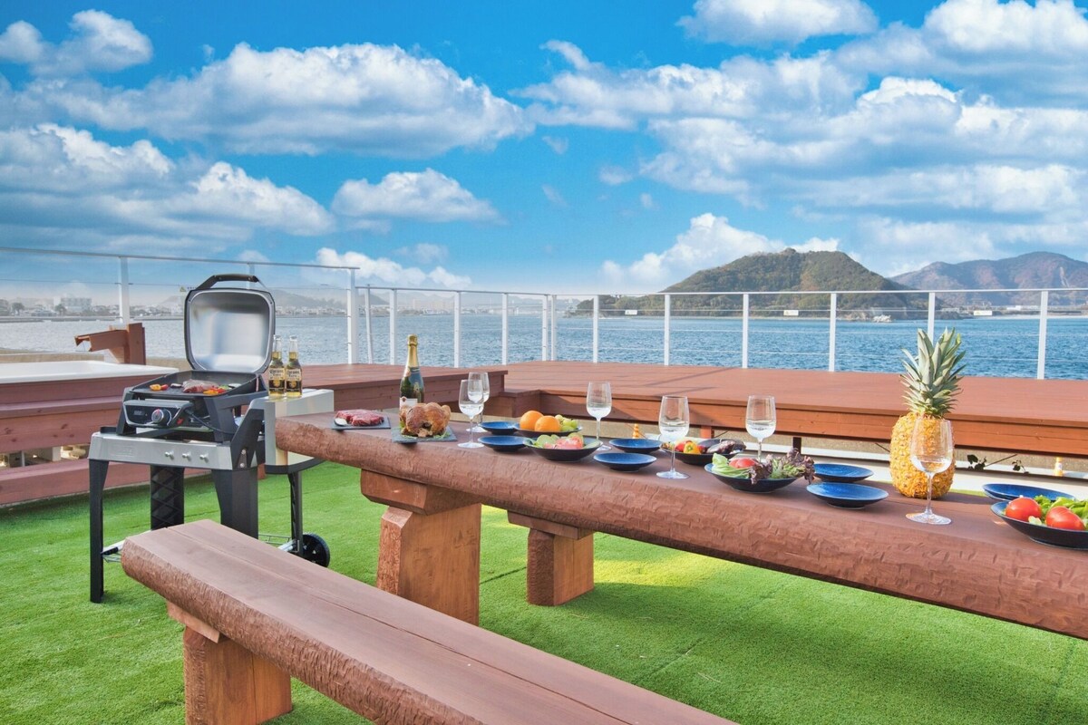 Un área de comedor al aire libre con una parrilla, bancos de madera y vistas al agua y las colinas.