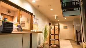 A reception area with a counter, a clock, a security camera, a signboard, a plant, a bookshelf, and a staircase. - Petit Hotel North Inn (Ito)