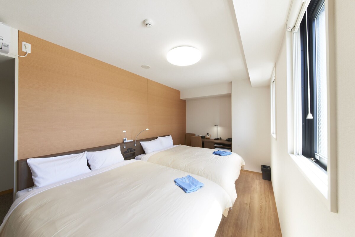 A hotel room with two beds, a desk, a chair, a window, and a ceiling light.