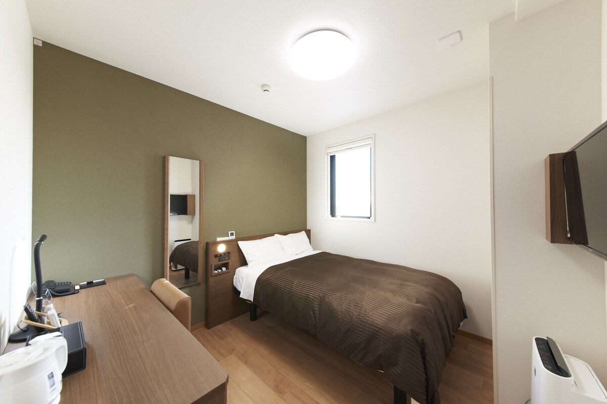 A hotel room with a bed, a desk, a chair, a TV, a window, and a mirror.