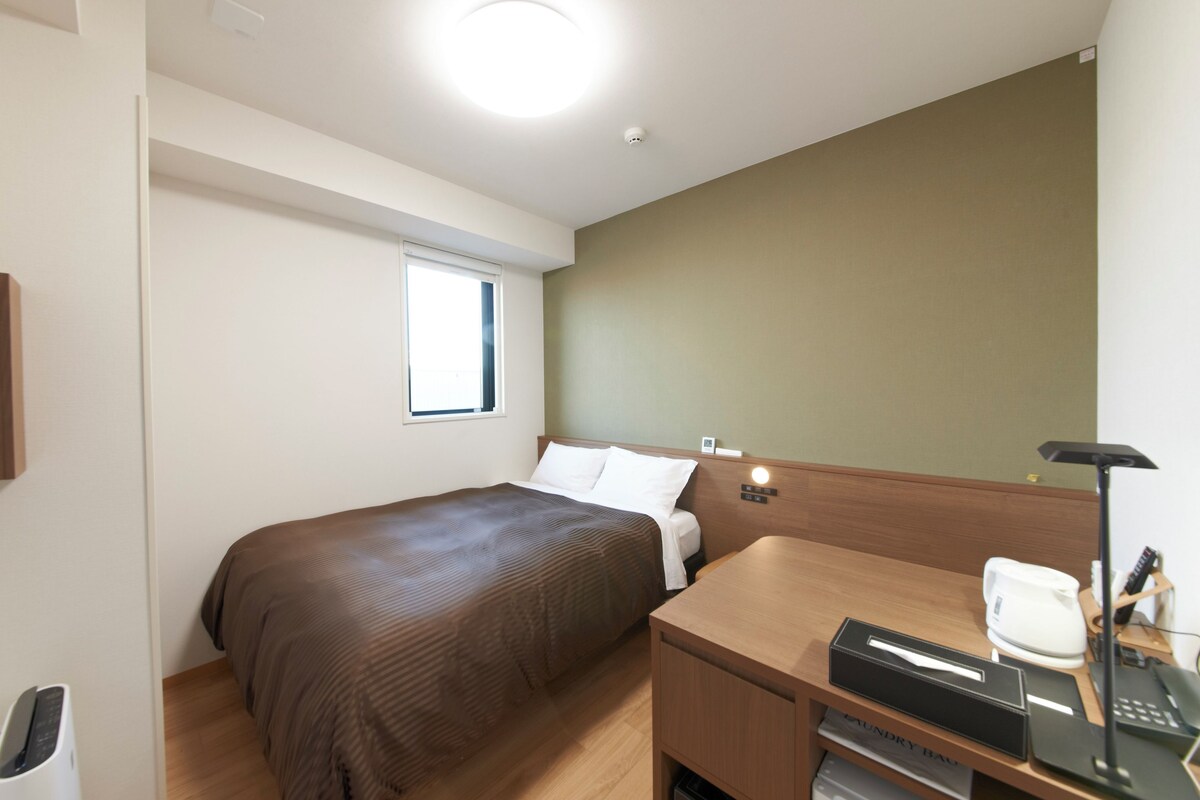 A hotel room with a bed, a desk with a telephone, a small window, and a wall-mounted light.
