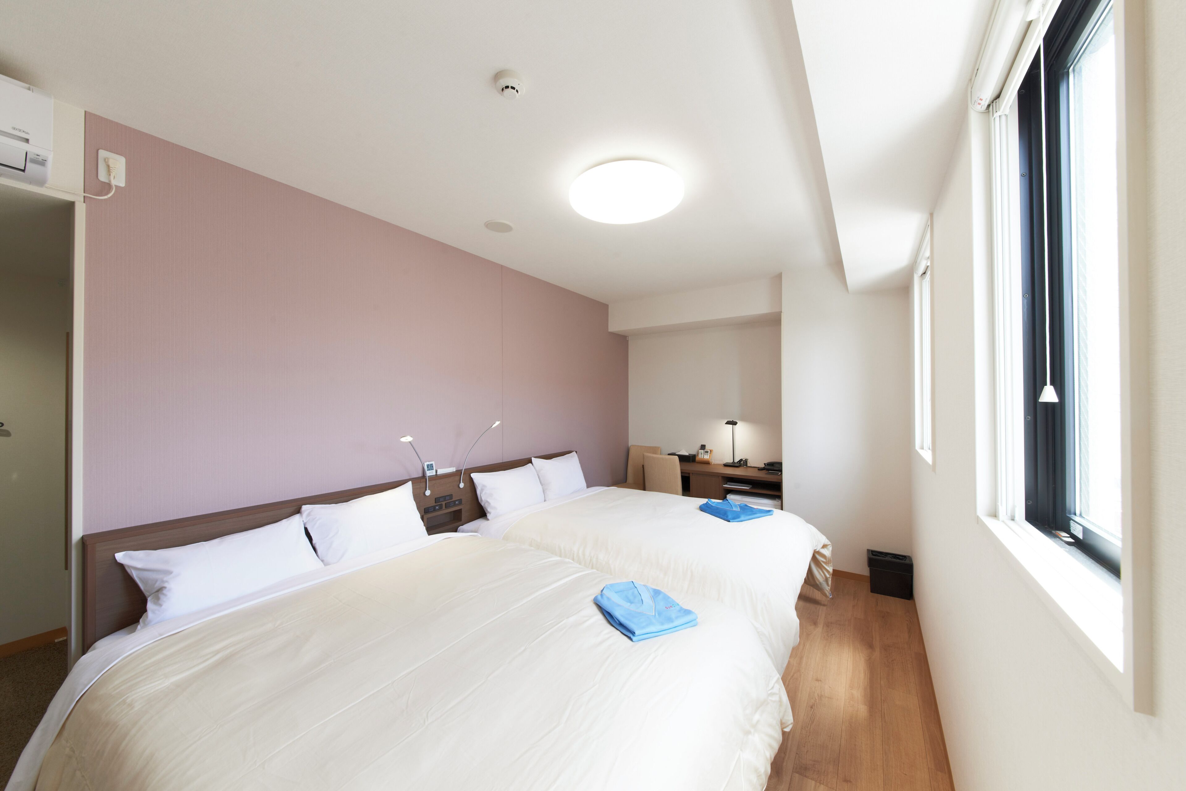 A hotel room with two beds, a desk, a chair, a window, and a ceiling light.