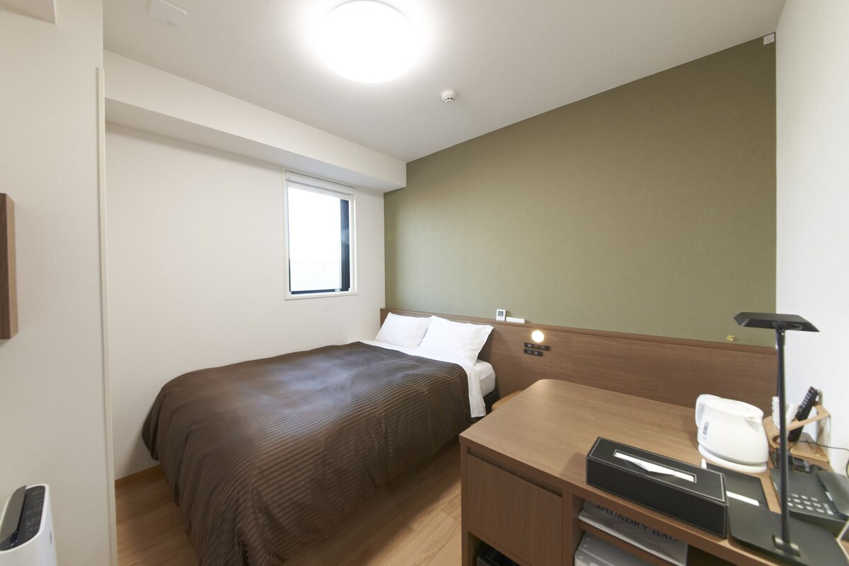 A hotel room with a bed, a desk, a chair, a window, and a bedside table with a lamp and a phone.