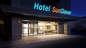 Entrada de un hotel con un cartel que dice 'Hotel SunClover', puertas de cristal y un área de descanso al aire libre.